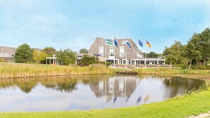 A building with multiple flags, including the European Union flag, a pond in front, and a grassy area. - North Sea Getaway – Hotel Near Dunes, Beach Trails & Ferry Port - 2 Units (Nes)