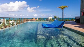 Piscina azul cristalina con una tumbona azul, rodeada de barandilla metálica y palmeras, con vista al océano y edificios al fondo.