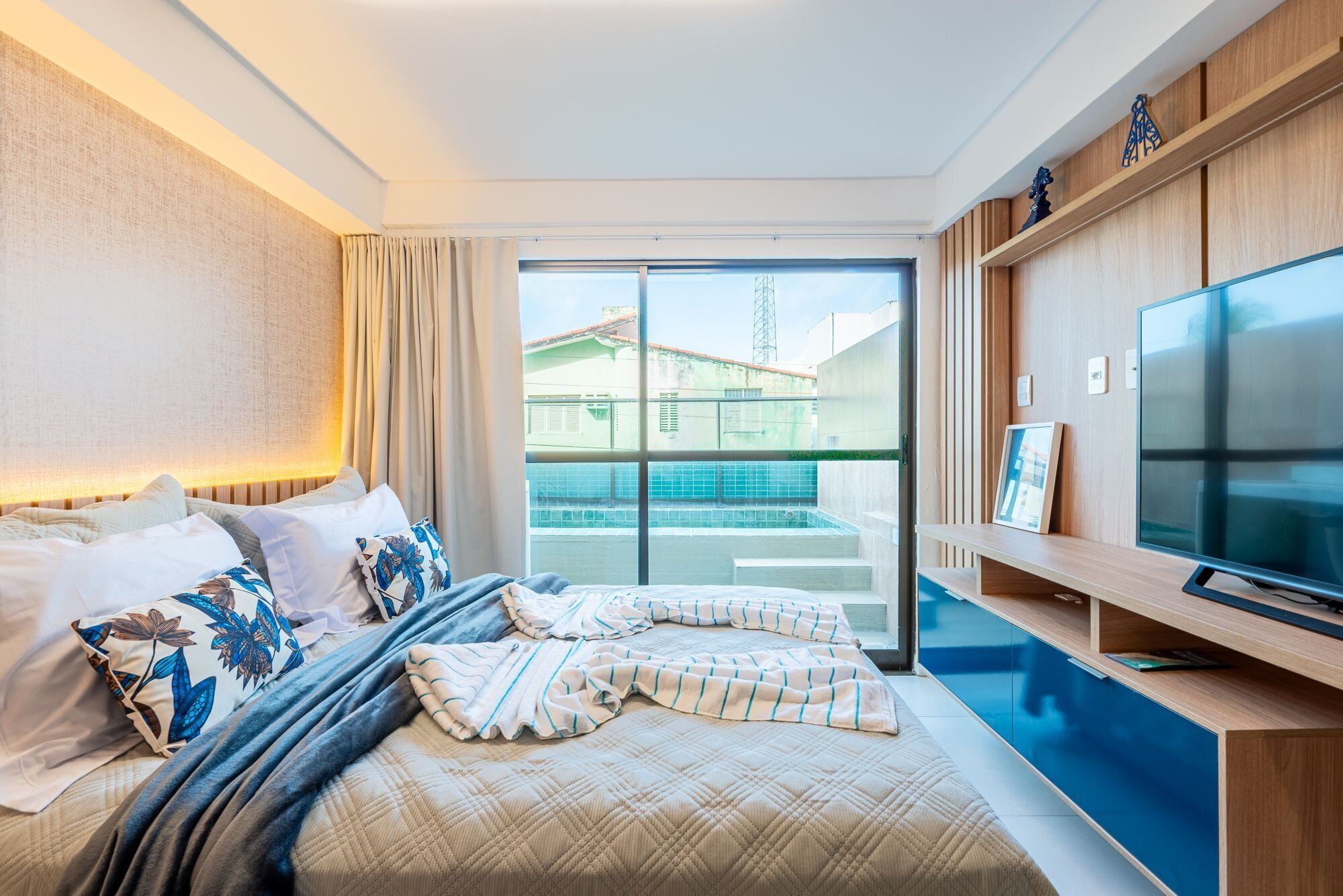 Habitación de hotel con cama, televisión, ventana y balcón con vista a edificios.