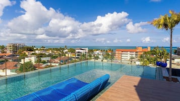 Una piscina azul cristalina con una tumbona azul, rodeada por una terraza de madera y palmeras, con vistas a un pueblo costero.