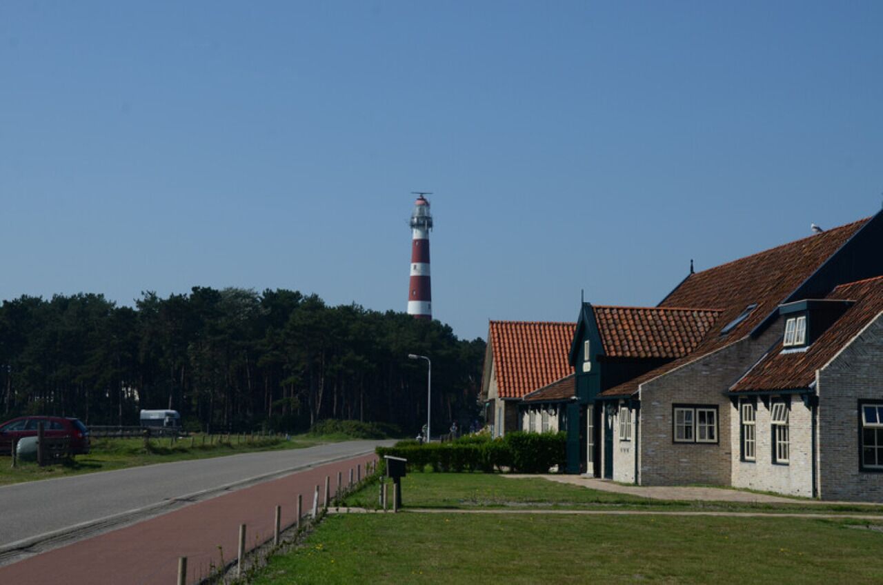 Un edificio costero con techo de tejas rojas, un faro al fondo y una carretera con carril bici rojo.