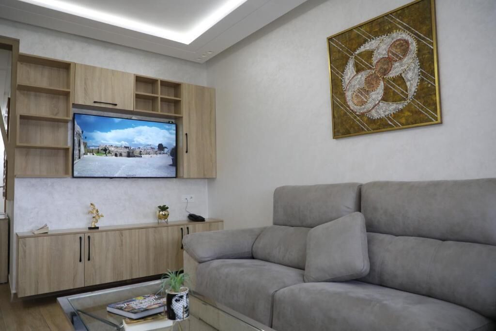 A modern living room with a flat-screen TV mounted on the wall, a grey sofa, and a coffee table with magazines and a plant.