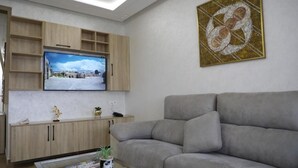 A modern living room with a flat-screen TV mounted on the wall, a grey sofa, and a coffee table with magazines and a plant.