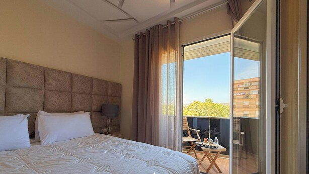 A hotel room with a large bed, a headboard, a sliding glass door leading to an outdoor balcony, and a view of the cityscape.
