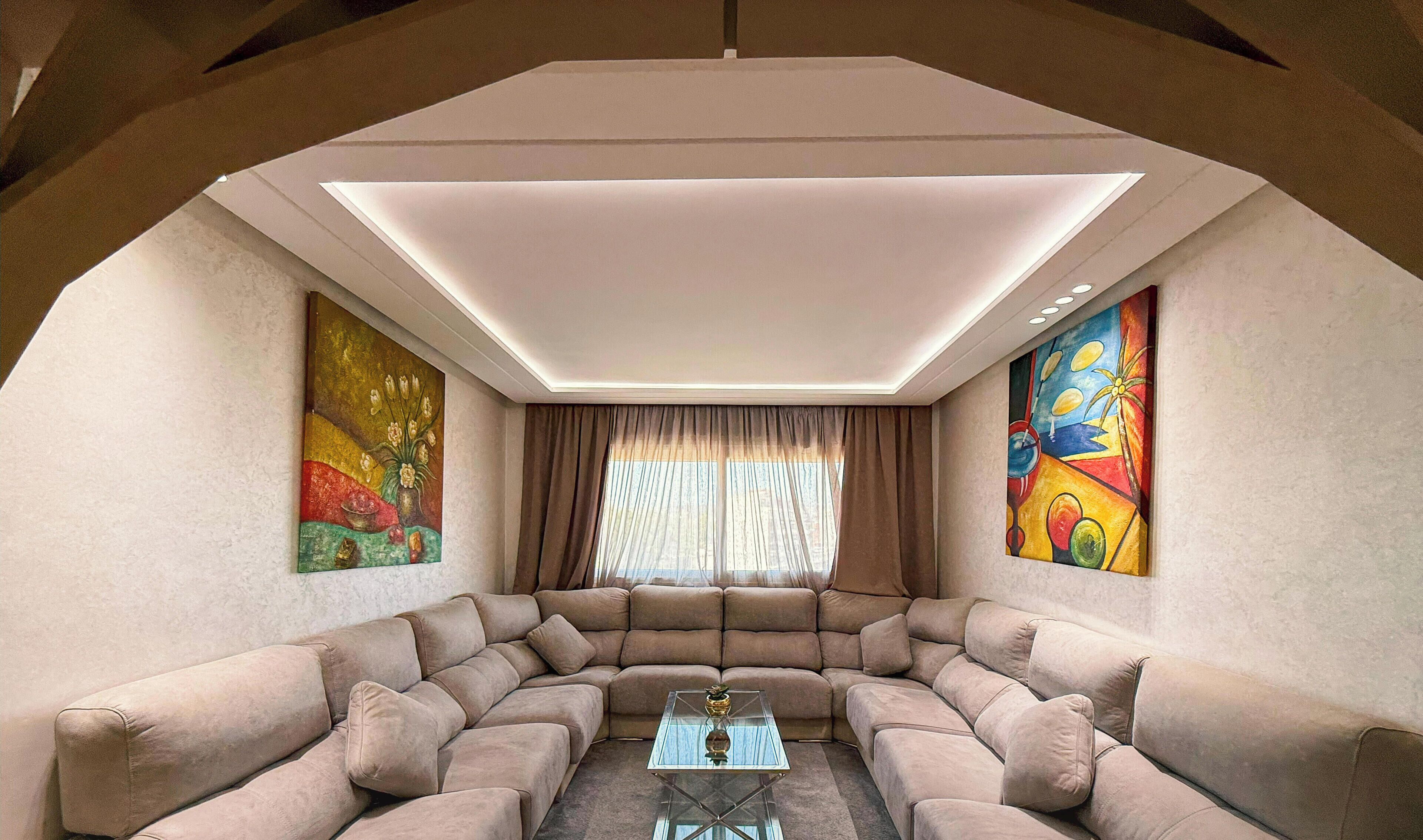A modern living room with a sectional sofa, a glass coffee table, and two large paintings on the walls.