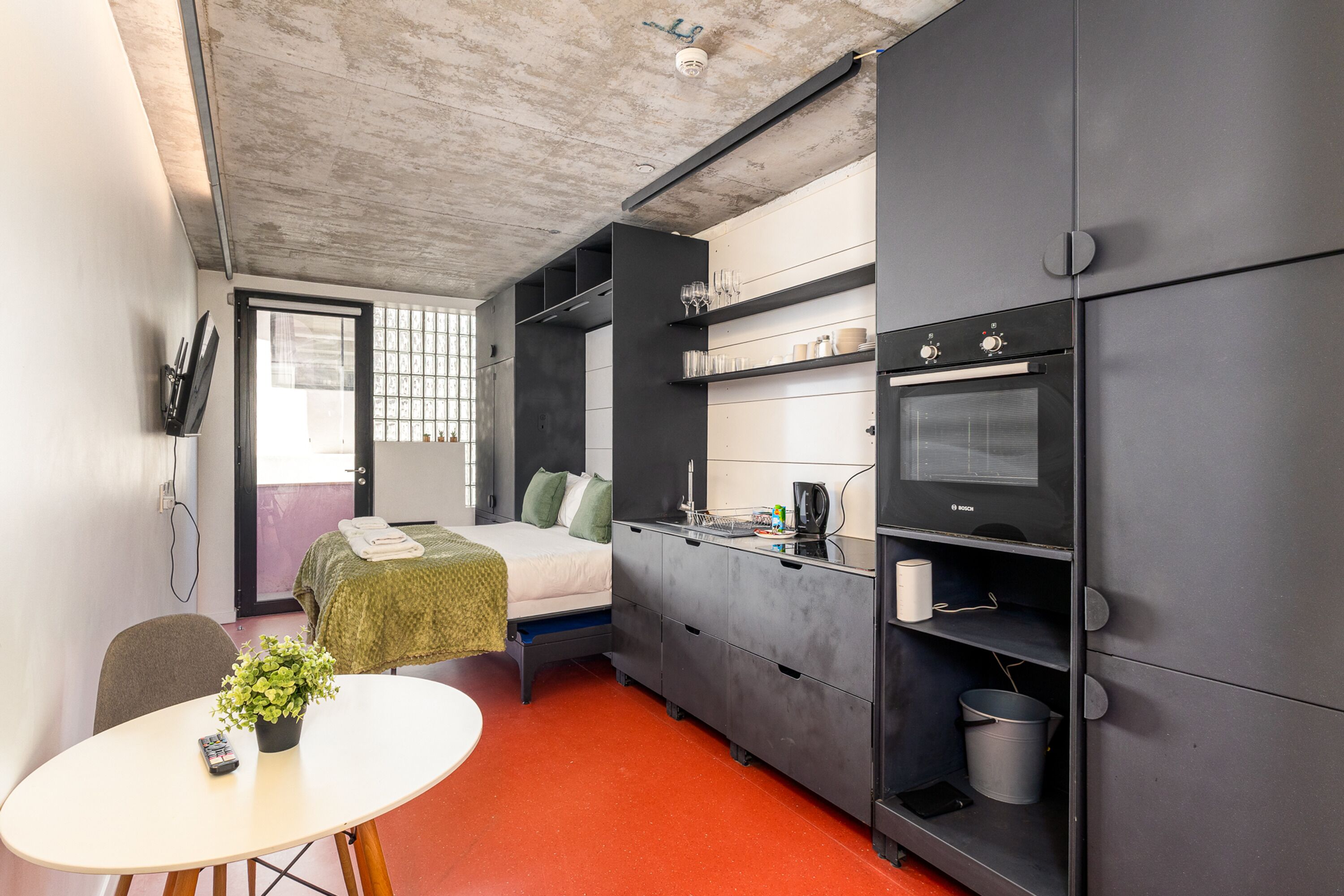 Habitación de hotel pequeña con cama, una mesita con una planta, kitchenette con horno Bosch y ventana con vistas.