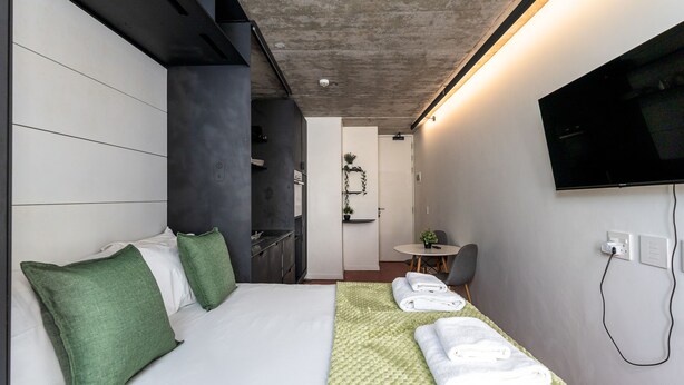 A modern hotel room with a bed, a TV mounted on the wall, a small table with a chair, and a bathroom visible through an open door. - The Urban Nest by Ctha (Cape Town)