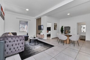 A modern living room with a sofa, a dining table with chairs, a TV mounted on the wall, and a window with blinds. - Luxury Living in Circa Illovo by Ctha (Johannesburg)