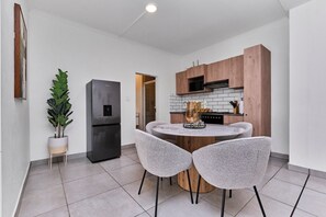 A compact kitchen with a central island, two chairs, a refrigerator, and wooden cabinets. - Luxury Living in Circa Illovo by Ctha (Johannesburg)