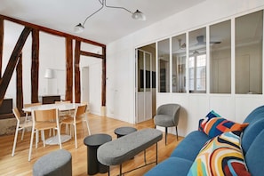 A modern living room with a dining area, wooden beams, large windows, and a blue sofa with colorful cushions. - A Stunning 2-BR in the 9th district- Folies (Paris)