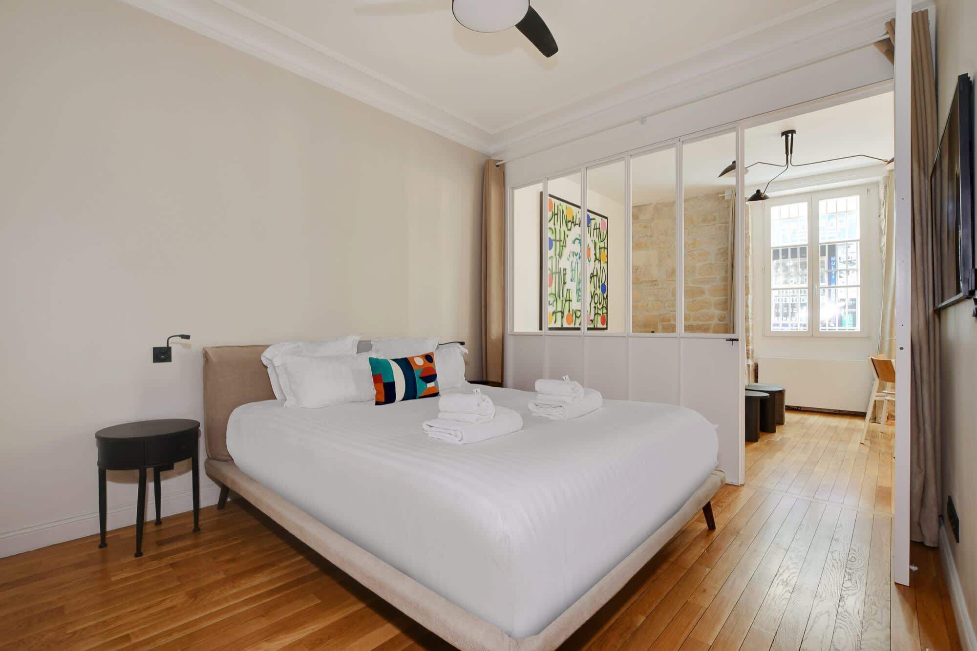 A modern bedroom with a large bed, a small side table, and a view of a cityscape through a window.