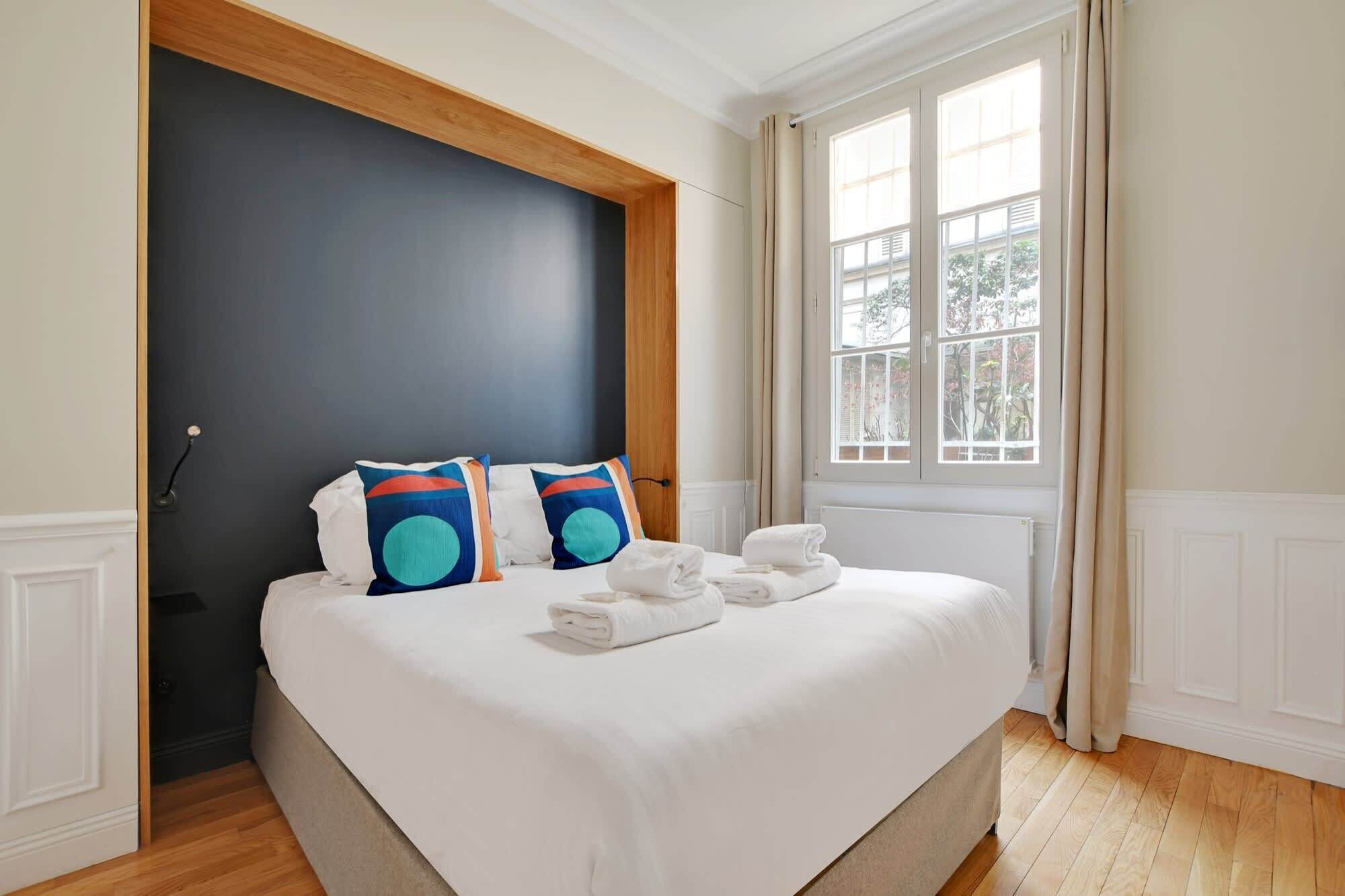A hotel room with a large bed, two pillows, a window with curtains, and a radiator.