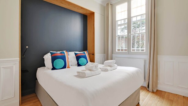 A hotel room with a large bed, two pillows, a window with curtains, and a radiator. - A Stunning 2-BR in the 9th district- Folies (Paris)