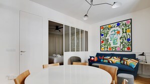 A modern living room with a round dining table, a sofa with colorful cushions, and a large abstract painting on the wall. - A Stunning 2-BR in the 9th district- Folies (Paris)
