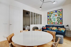 A modern living room with a round dining table, a sofa with colorful cushions, and a large abstract painting on the wall. - A Stunning 2-BR in the 9th district- Folies (Paris)