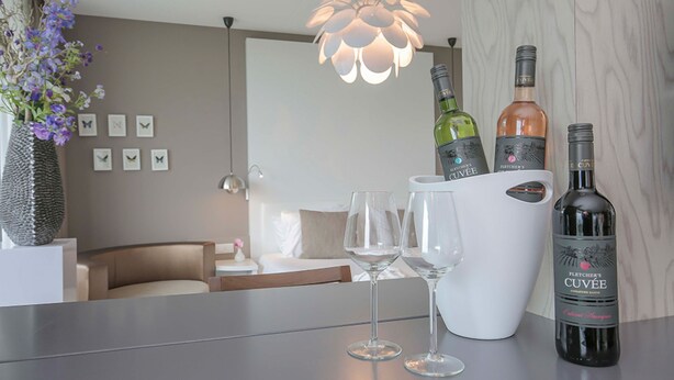 A hotel room with a bed, a bedside table with two wine glasses, a vase with flowers, and a bottle of Fletcher's Cuvée on the table. - Spacious Family Stay | Wellness Access & Outdoor Adventure | Valley, Forest Site (Wageningen)