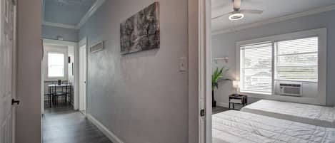 A hallway leading to a bedroom with a ceiling fan, a window with blinds, and a painting on the wall.