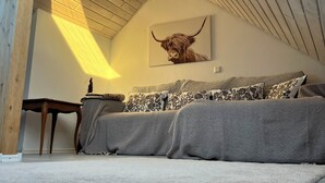 A room with a sloped ceiling, a sofa with patterned cushions, a wall-mounted bull head artwork, and a small table with a lamp. - Familienurlaub nahe Europa-Park & Rulantica mit Garten, Trampolin & Balkon (Herbolzheim)