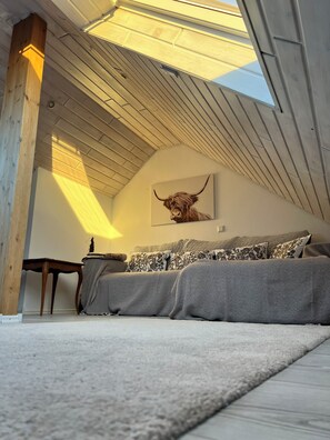 A room with a sloped ceiling, a sofa with patterned cushions, a wall-mounted bull head artwork, and a small table with a lamp. - Familienurlaub nahe Europa-Park & Rulantica mit Garten, Trampolin & Balkon (Herbolzheim)