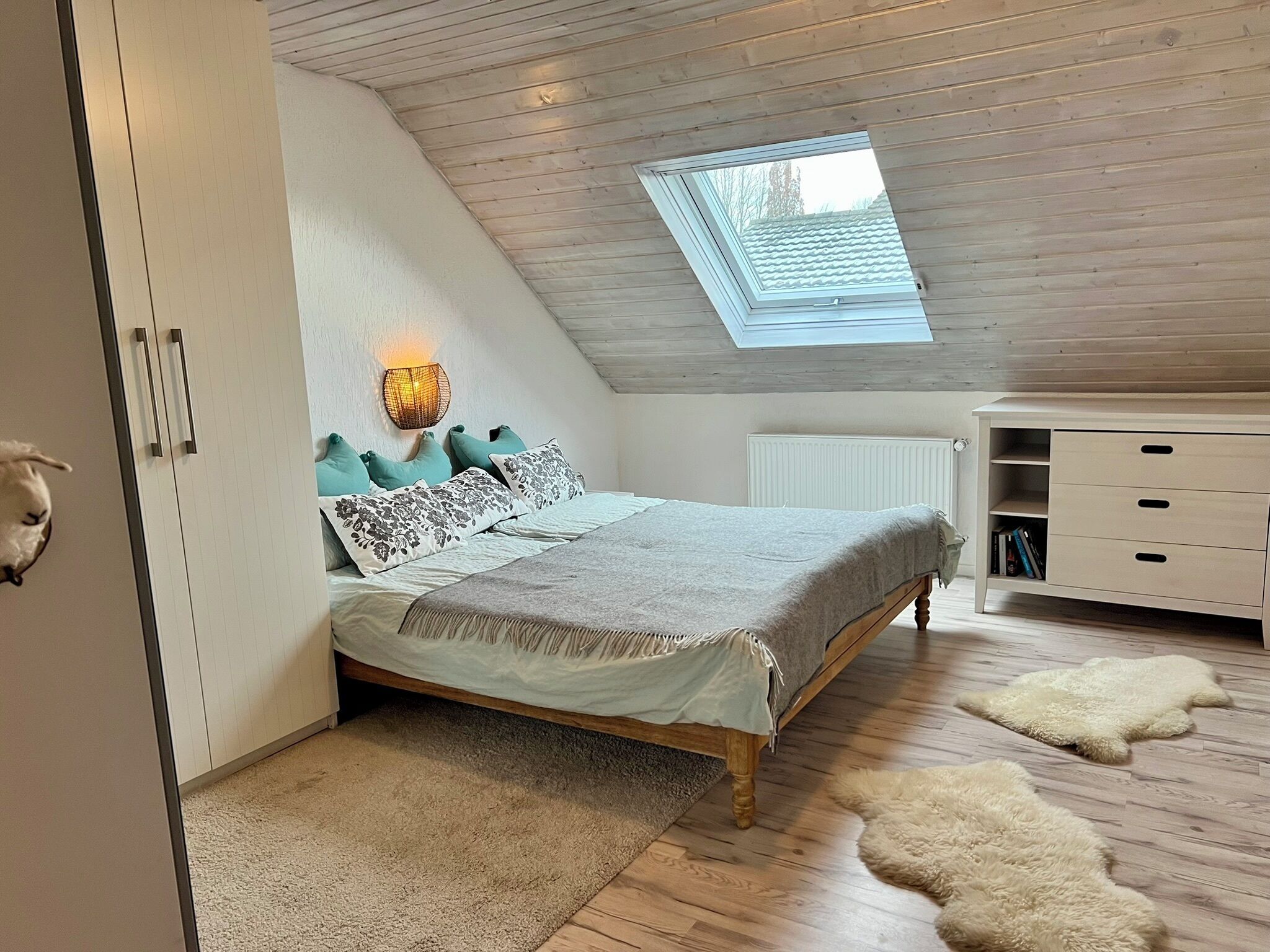 Ein Schlafzimmer mit schrägen Wänden, einem Oberlicht, einem Bett mit gemusterten Textilien, einer Nachttischlampe, einer Heizung und einer weißen Kommode.