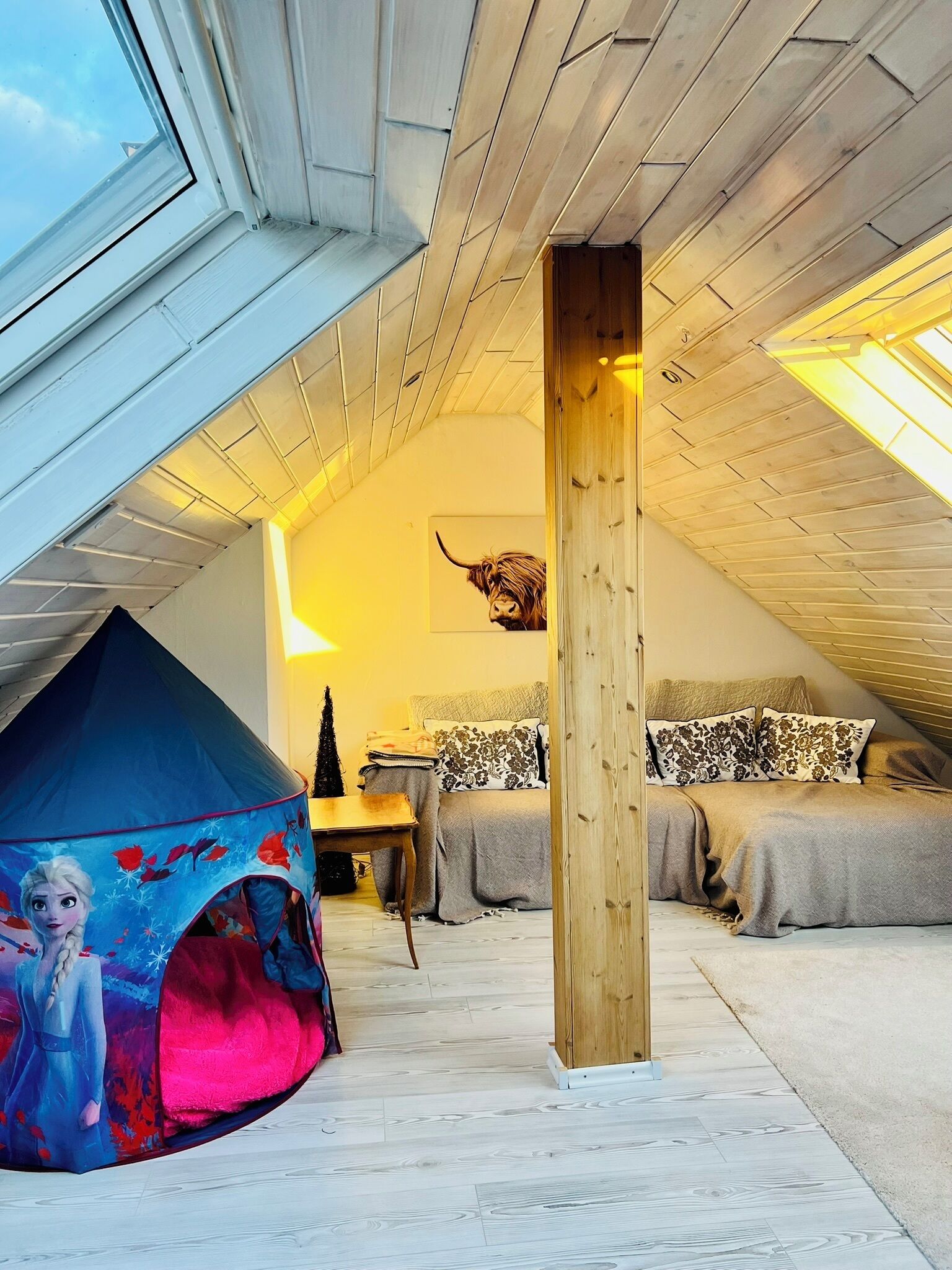 Ein Schlafzimmer mit schräg verlaufender Decke, einer zeltartigen Konstruktion, einem Holzbalken und einem Bett mit gemusterten Kissen.