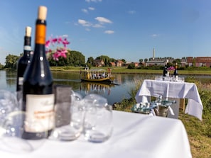 A lakeside table set with wine bottles and glasses, a boat in the background, and a building with a chimney. - Family Friendly Nature Escape w/Dining & Spa | Nature Reserves & Local Landmarks (Wageningen)