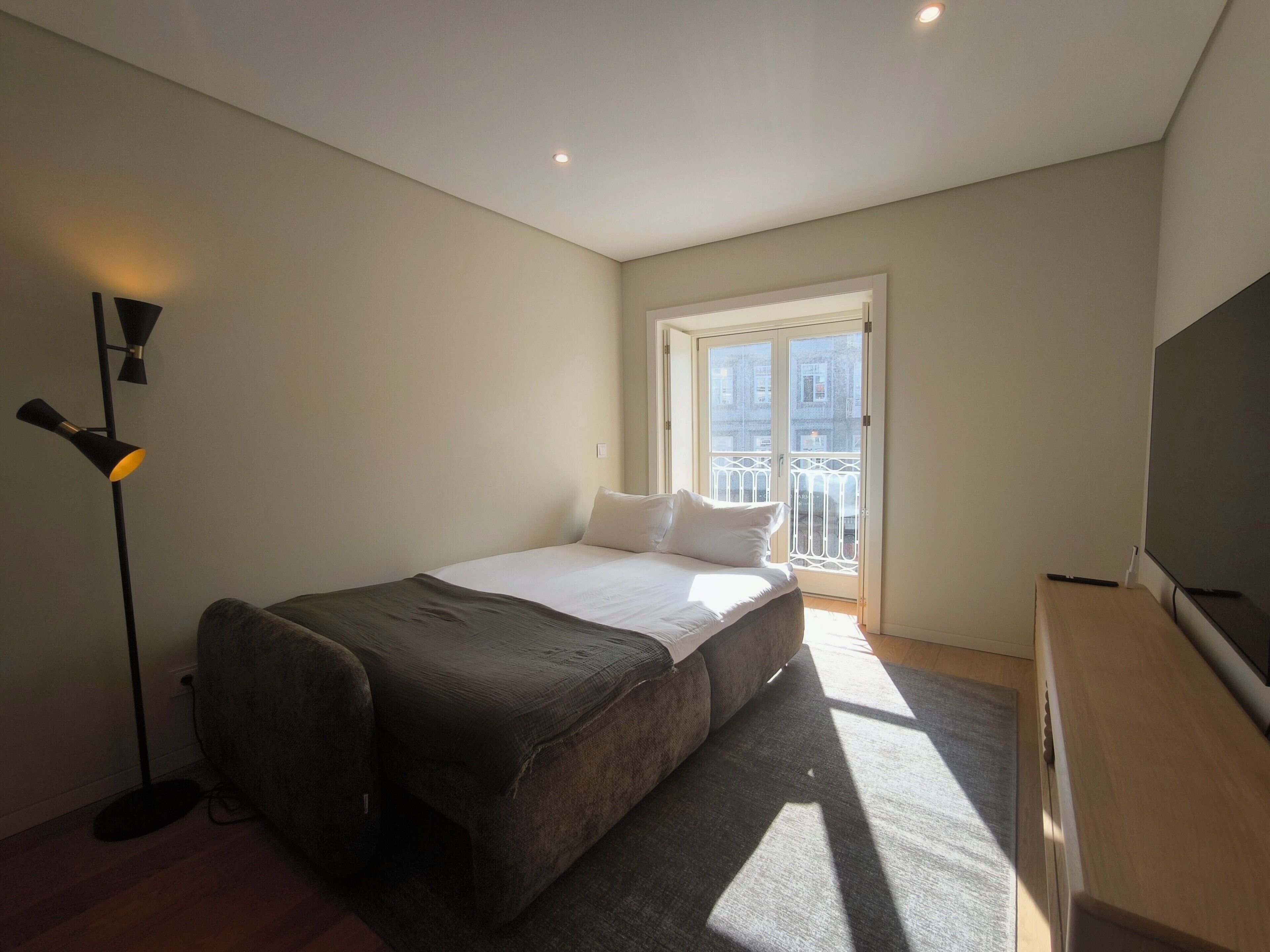 A bedroom with a bed, a desk, a floor lamp, and a window with a view of buildings.