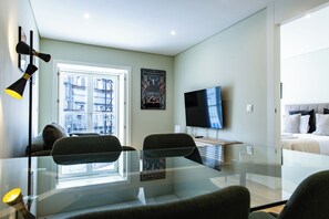 A modern hotel room with a glass dining table, two chairs, a flat-screen TV, a poster on the wall, and a bed with pillows. - Carmo Cinema Apartments (Porto)