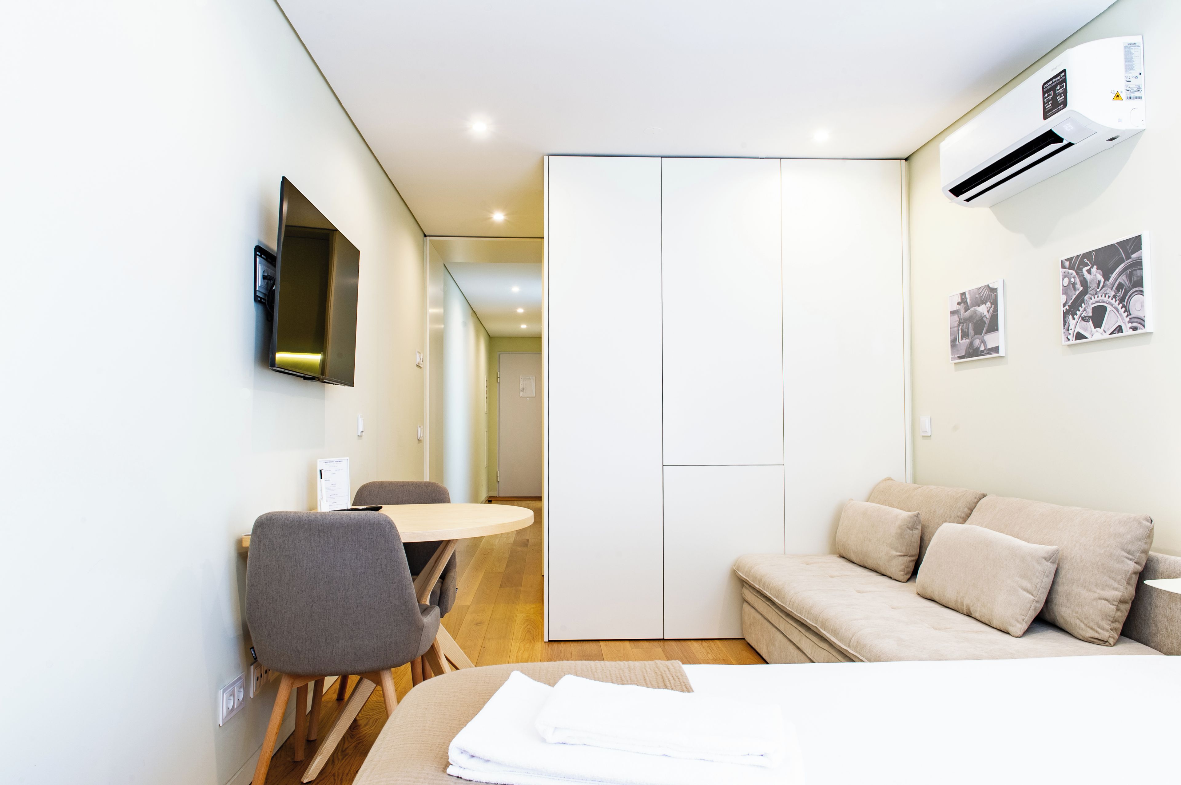 A modern hotel room with a bed, a desk, a chair, a flat-screen TV mounted on the wall, and an air conditioning unit.