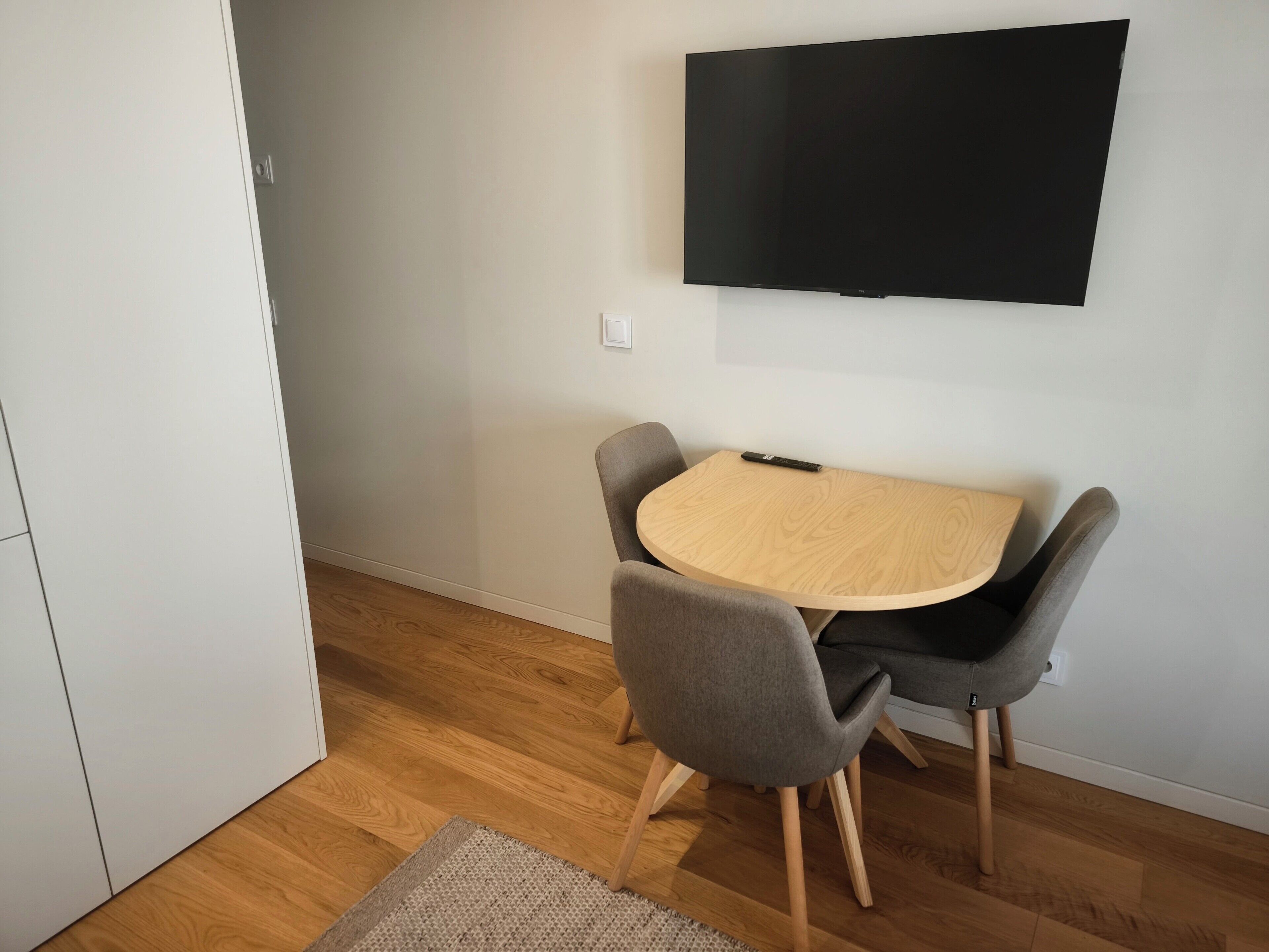 Una habitación con una mesa de madera, dos sillas grises, un televisor de pantalla plana montado en la pared y suelo de madera.