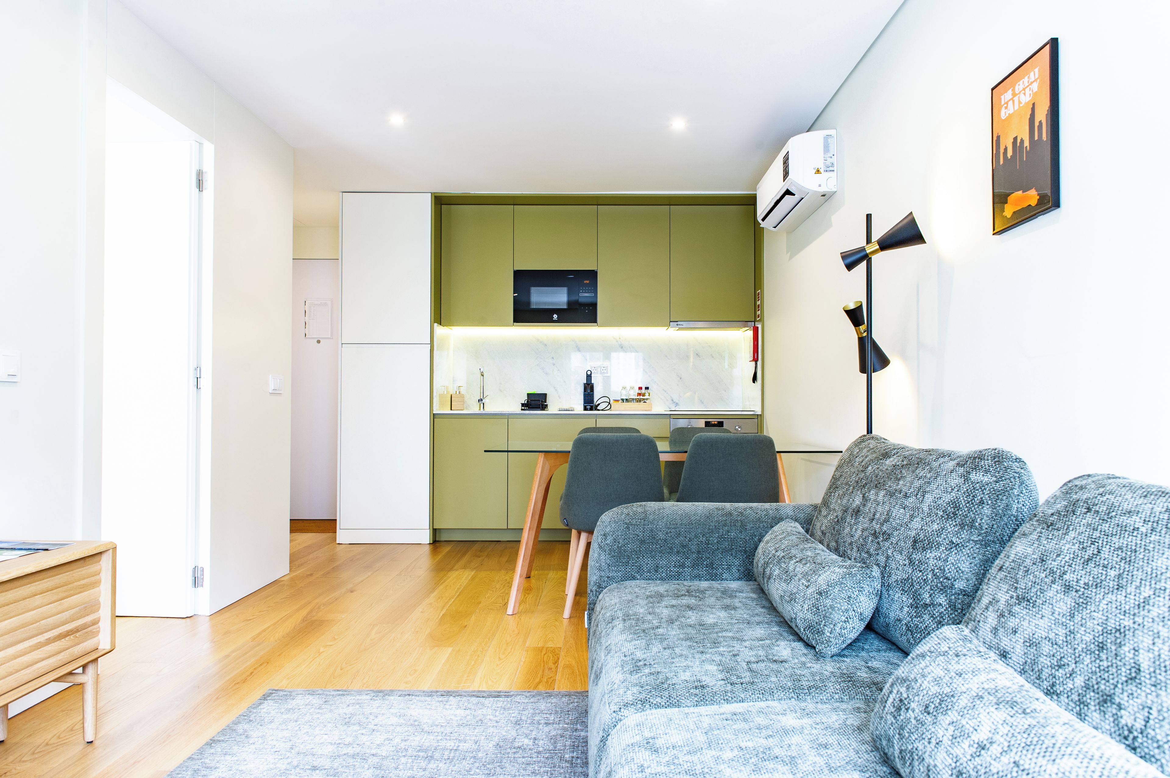 Una sala moderna con un sofá gris y un banco de madera, una cocina con armarios verdes y un microondas, además de un cartel enmarcado en la pared.
