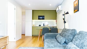 Uma sala de estar moderna com um sofá cinzento e um banco de madeira, uma cozinha com armários verdes e um micro-ondas, e um pôster emoldurado na parede.
