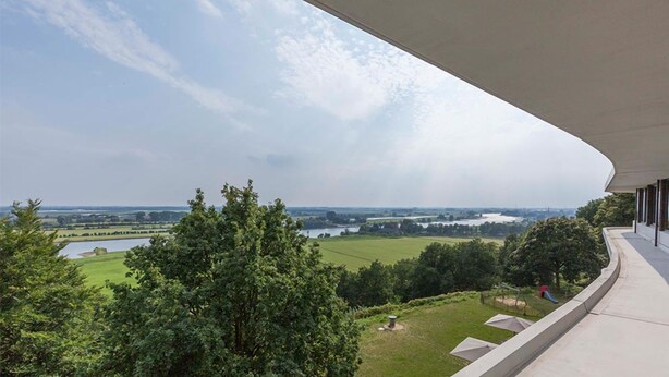 Un edificio moderno con un tetto curvo che si affaccia su un paesaggio verde e uno specchio d'acqua.