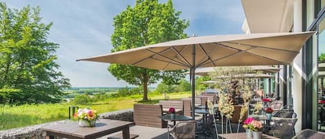 An outdoor patio with tables and chairs, a large umbrella, and a view of the landscape.