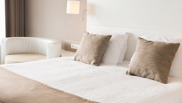 Quarto de hotel com cama, duas almofadas, cadeira, mesa de cabeceira e luminária.