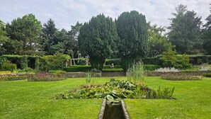 Um jardim com um lago central, rodeado por relvado bem cuidado e diversas plantas.
