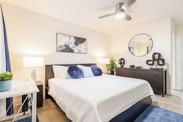 Ein modernes Schlafzimmer mit Bett, Nachttischen, Schreibtisch, Kommode, Deckenventilator, Spiegel und einem Wandbild.