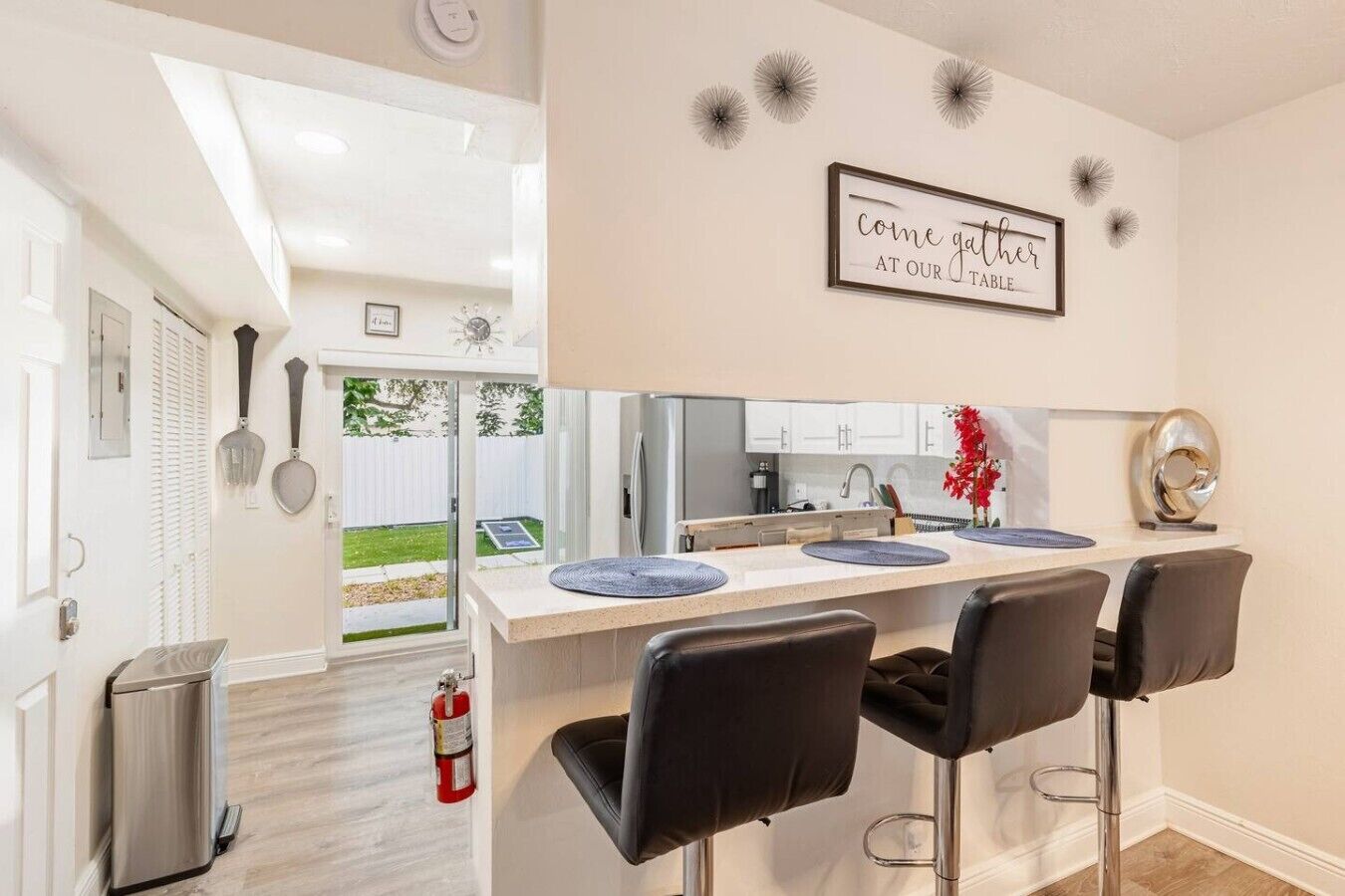 A modern kitchen with a central island, bar stools, a fire extinguisher, and a sign inviting guests to the table.