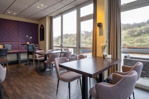 Uma sala de jantar moderna com mesas e cadeiras, um espaço de estar com um banco acolchoado e grandes janelas com vista para o exterior.