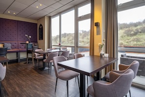 Uma sala de jantar moderna com mesas e cadeiras, um espaço de estar com um banco acolchoado e grandes janelas com vista para o exterior.