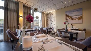 Een restaurantgedeelte van een hotel met gedekte tafels, een schilderij aan de muur en uitzicht op het omliggende landschap.