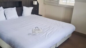 Una cama bien hecha con sábanas blancas, una mesita de noche con lámpara y una ventana con persianas.