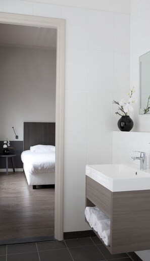 A modern hotel room with a large bed, a bedside table, a mirror, and a bathroom with a sink and towel rack.