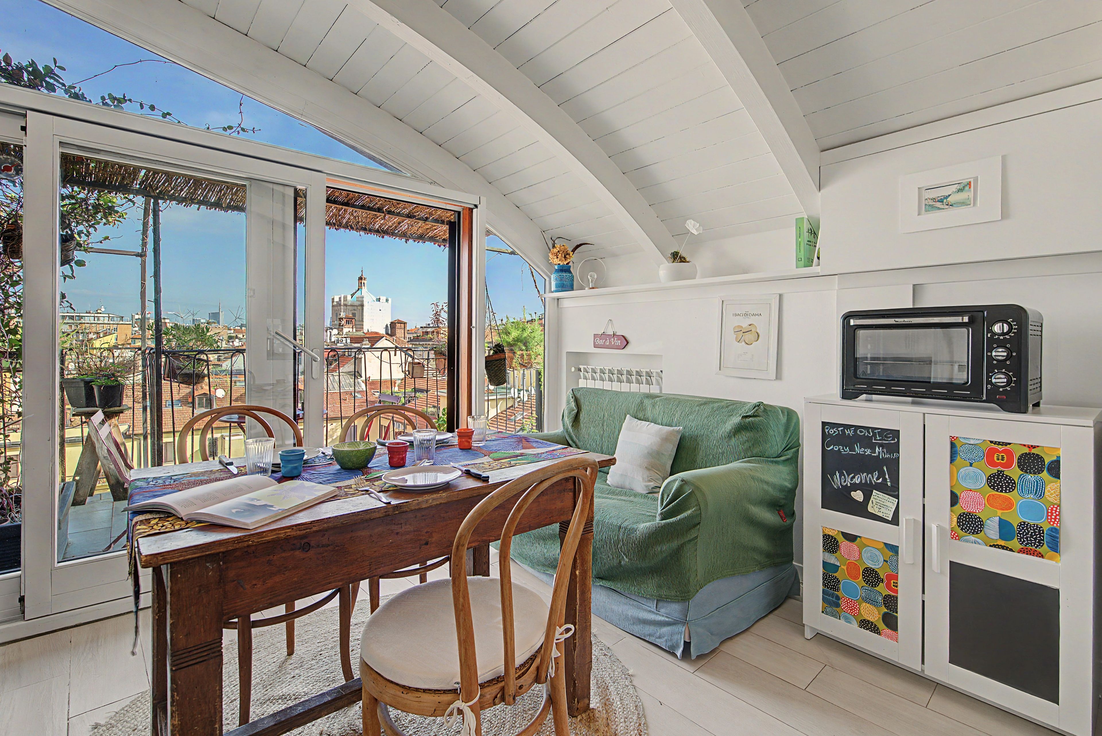 A dining area with a table set for two, a green sofa, a white cabinet with a chalkboard, and a view of buildings through large windows.