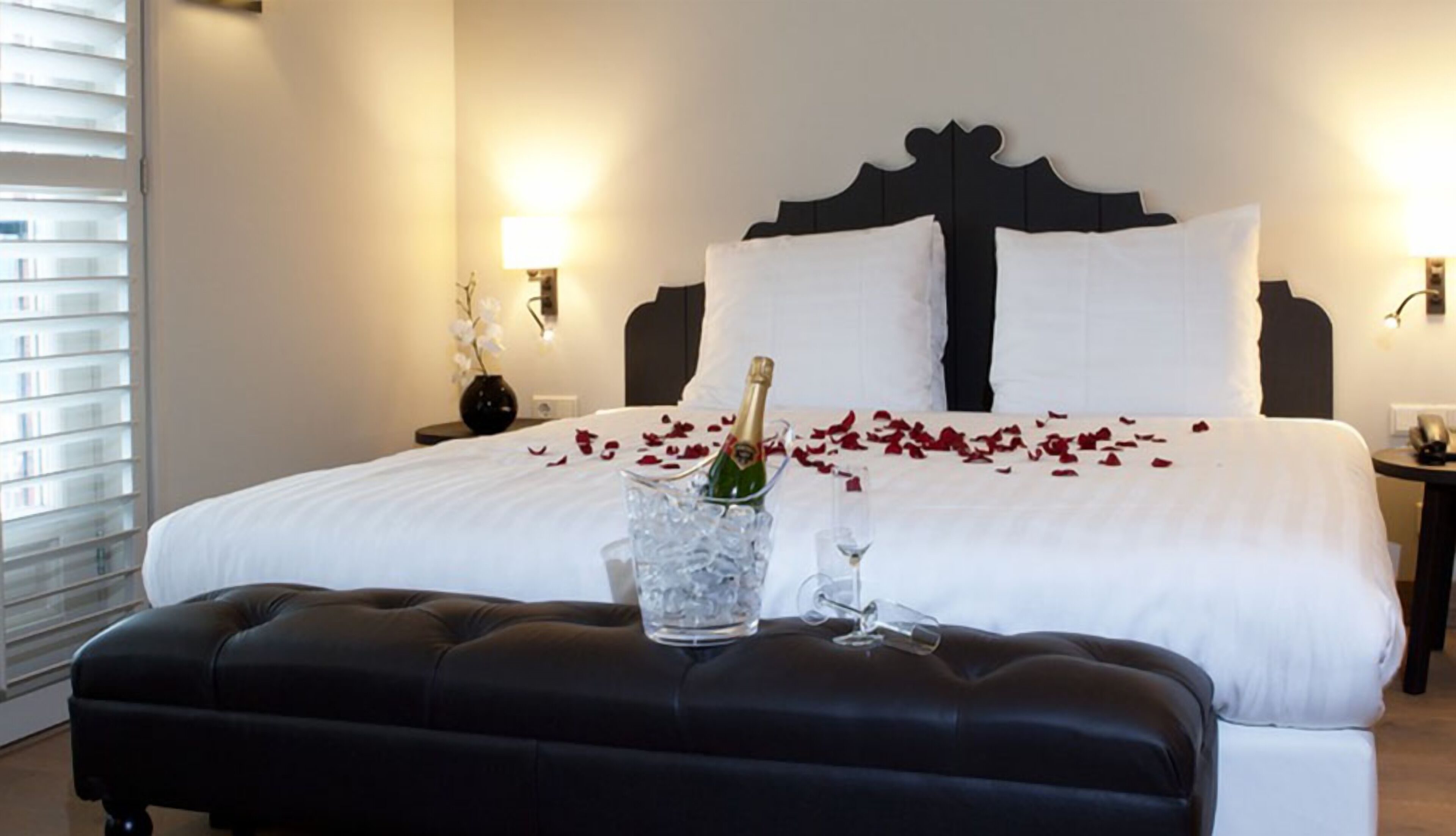 Een hotelkamer met een groot bed, een nachtkastje met een bloemenvaas en een fles champagne op het bed.