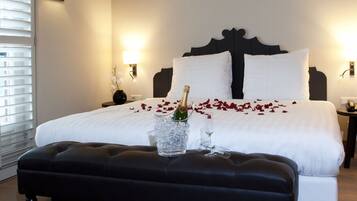 Een hotelkamer met een groot bed, een nachtkastje met een bloemenvaas en een fles champagne op het bed.