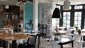 Interior de restaurante com várias mesas e cadeiras, algumas com plantas. Há luminárias pendentes e uma grande janela com vista.