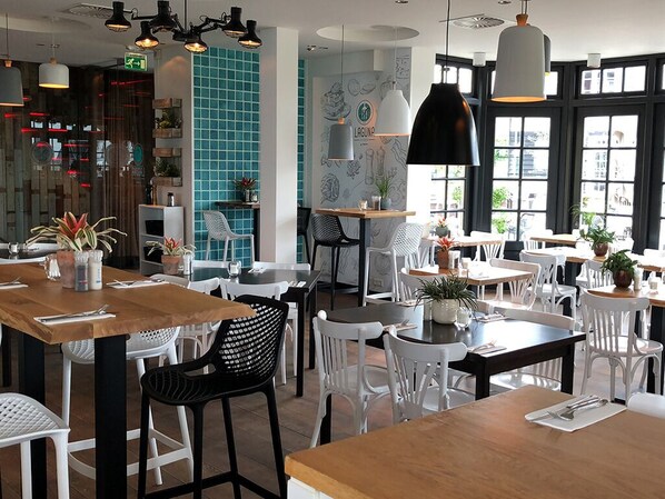 A restaurant interior with multiple tables and chairs, some with plants. There are hanging lights and a large window with a view.