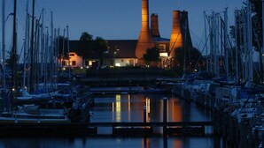 Un port de plaisance avec des bateaux amarrés, un bâtiment doté de deux hautes cheminées et un pont au premier plan.