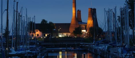 A marina with boats docked, a building with two tall chimneys, and a bridge in the foreground.